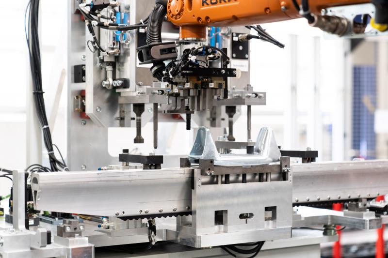Der Roboter legt den Kettensteg in die erste Montagestation. Anschließend legt der kleine Roboter eine Zwischenlage aus Kunststoff auf, die verhindern soll, dass zwischen Aluminiumprofil und Stahlbügel Kontaktkorrosion auftritt. Danach wird ein sogenannter Stahlgussbügel aufgeschraubt, wobei die Verschraubung kontrolliert und dokumentiert wird. In weiteren Prozessschritten wird der Kettensteg mit Eisgreifern vervollständigt